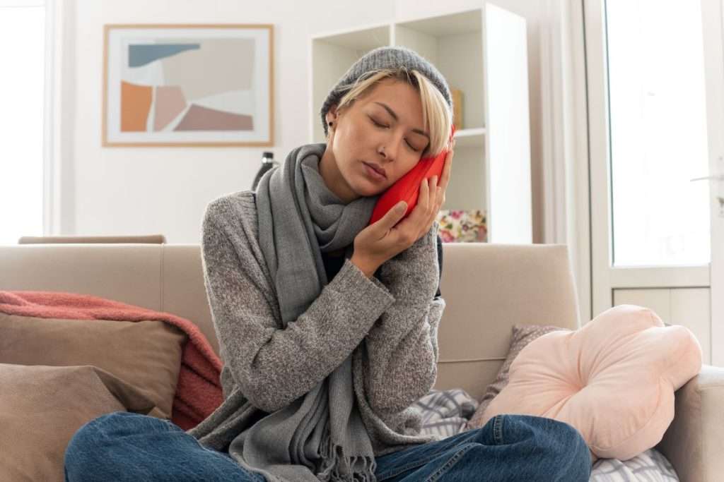 Young woman sitting on couch applying cold compress to her cheek after wisdom tooth removal for pain relief.
